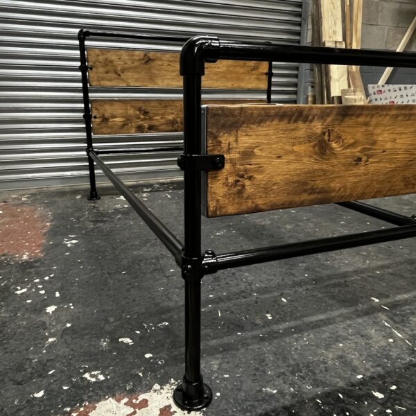 Industrial-style metal and wood bed frame with slatted wooden base in a workshop setting, featuring black metal pipes and wooden panels on the headboard and footboard
